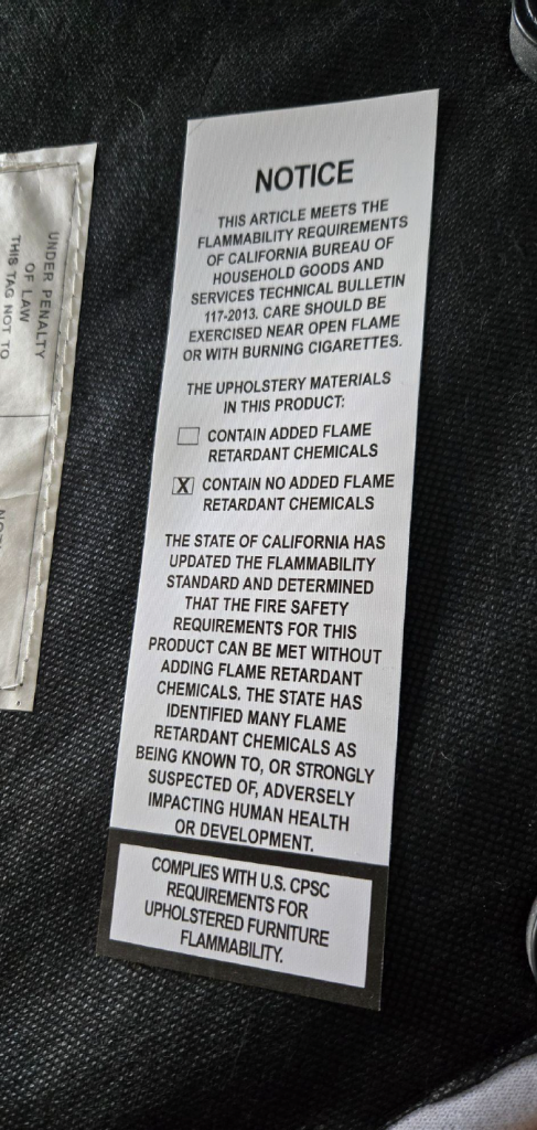 Close-up photo of a TB-117-2013 compliant flammability label showing required size, permanent attachment, flame-retardant disclosure checkboxes, and CPSC certification statement.